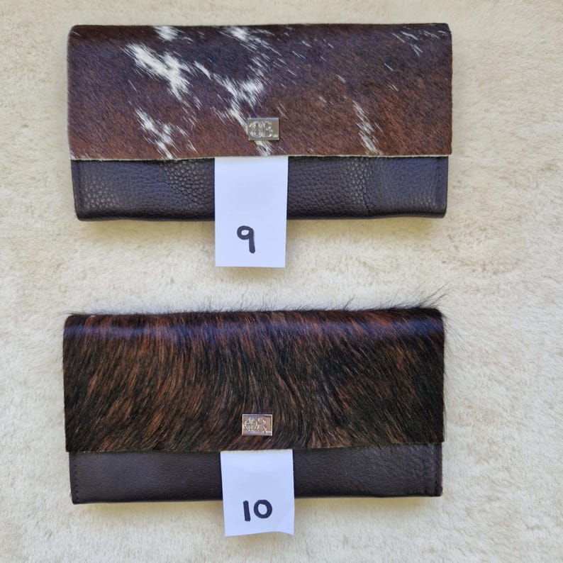 May include: Two leather wallets with a cowhide flap. One wallet has a brown and white cowhide flap, and the other has a brown cowhide flap. Both wallets have a dark brown leather base and a silver metal clasp. The number "9" and "10" are on the wallets.