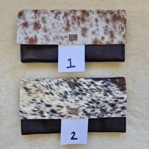 May include: Two leather wallets with cowhide flap closures. One wallet has a brown and white spotted pattern, and the other has a black and white spotted pattern. Both wallets have a dark brown leather base and a silver-colored clasp.