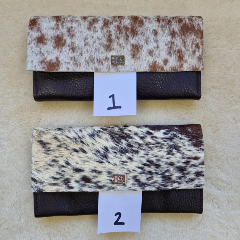 May include: Two leather wallets with cowhide flap closures. One wallet has a brown and white spotted pattern, and the other has a black and white spotted pattern. Both wallets have a dark brown leather base and a silver-colored clasp.