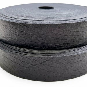 May include: A roll of black ribbon featuring a textured crocodile skin pattern. The material has a matte finish and is wound into a tight, circular spool. The surface of the ribbon displays a repeating grid-like embossed design throughout.
