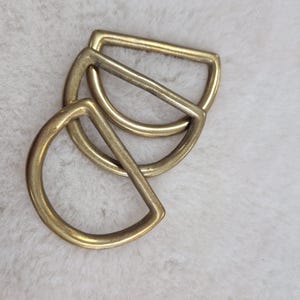 May include: Three brass-coloured D-rings are arranged on a white, textured surface. The D-rings are made of metal and have a rounded shape on one side and a straight bar on the other. The rings are slightly overlapping, suggesting they are for crafting or hardware.