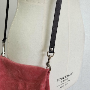 May include: A close-up of a dusty rose suede crossbody bag with a black leather strap and silver-toned hardware. The bag is attached to a black leather strap with a silver clasp. The background is a mannequin.