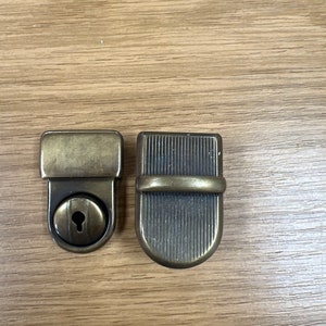 May include: Two antique brass-toned metal clasps. One is a rectangular lock with a keyhole, and the other is a matching clasp with vertical lines and a curved bottom. Both are set against a light brown wooden surface.