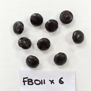 May include: A collection of eight round, dark brown buttons with a woven leather-like texture and a smooth finish. Each button features a cross-stitch pattern on the surface. A white paper label at the bottom displays the handwritten text FB011 x 6.