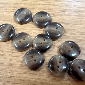May include: A collection of ten brown and tan buttons with two holes each. The buttons have a slightly irregular, square shape and a glossy finish. The buttons are scattered on a light brown wooden surface.