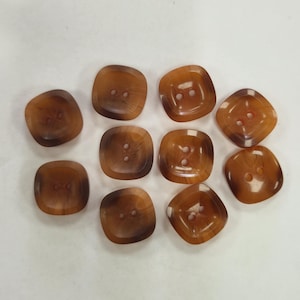 Puede incluir: Una colección de diez botones cuadrados marrones con esquinas redondeadas. Cada botón tiene dos agujeros para coser y un aspecto veteado, con bordes más oscuros y centros más claros. Los botones están dispersos sobre una superficie blanca.
