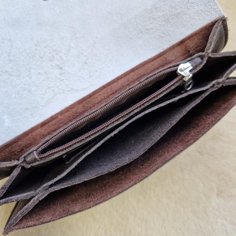 May include: Open view of a brown leather wallet with multiple compartments. The wallet has a zipper closure and a light-colored interior. The leather appears to be distressed, giving it a rustic look. The wallet is designed to hold cards, cash, and other essentials.