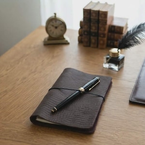May include: A dark brown textured notebook with a black elastic band closure and a black fountain pen resting on top. Nearby are a small gold-toned analog clock, a stack of vintage books, a glass inkwell with a black feather quill, and a leather pad.