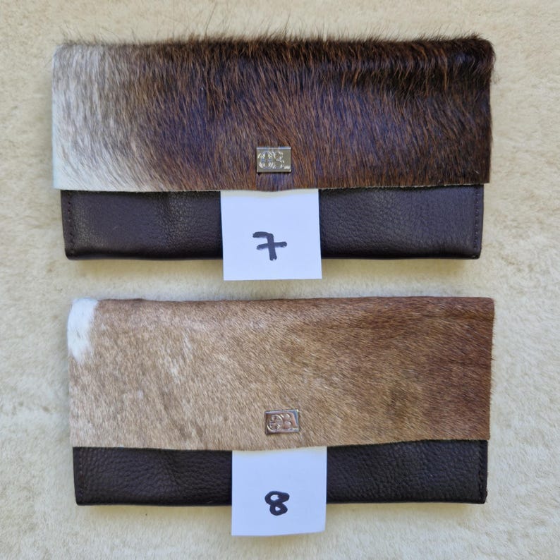 May include: Two leather wallets with a flap closure. The top portion of the wallets is covered in brown and white animal hide. The bottom portion is dark brown leather. Each wallet has a silver metal emblem and a white tag with a number.