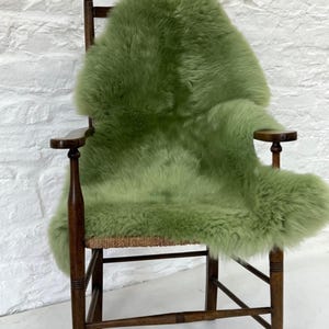May include: A wooden armchair with a woven seat and a green sheepskin throw. The sheepskin is a soft, fluffy texture and the chair has a dark wood finish. The background is a white brick wall.