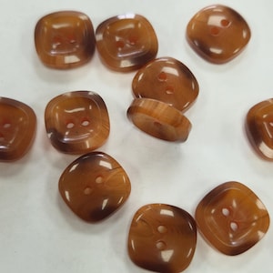 May include: A selection of brown, square buttons with rounded corners. Each button features two sewing holes. The buttons have a marbled effect, with varying shades of brown throughout.
