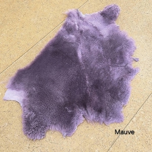 May include: A mauve-coloured sheepskin pelt with a thick, soft, and dense wool texture. The hide features an irregular, natural shape with a plush surface and a consistent purple-toned colour throughout. The word Mauve is printed in black text.
