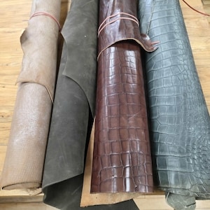 May include: Four rolls of leather material arranged side by side. The first roll is light tan with a fine texture, the second is matte dark grey, the third is glossy dark brown with a crocodile skin pattern, and the fourth is grey with a crocodile pattern.