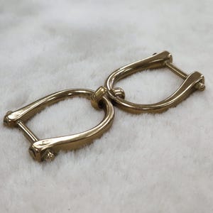May include: A pair of gold-colored metal D-ring connectors, linked together. Each D-ring has a curved shape with a straight bar and a small screw-like fastener. The connectors are on a soft, white surface.
