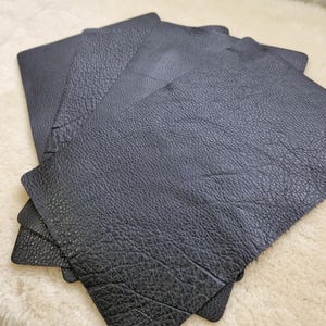 May include: Several black leather pieces are stacked, showing a textured surface. The leather pieces are rectangular with rounded corners. The leather has a slightly rough, grainy appearance.