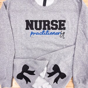 Könnte beinhalten: Grauer Pullover mit schwarzem Sticktext, der "NURSE practitioner" mit einem blauen Stethoskop-Symbol lautet. Der Pullover hat schwarze gestickte Schleifen an den Ärmeln.