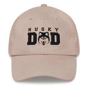 May include: Tan colored baseball cap with black embroidered text that reads "HUSKY DAD" and a black and white husky silhouette.