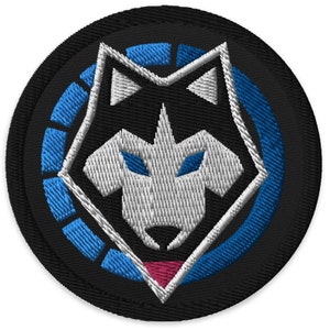 May include: A black and blue embroidered patch with a white wolf head design. The wolf has blue eyes and a pink nose. The wolf is surrounded by a blue circle with a white outline.