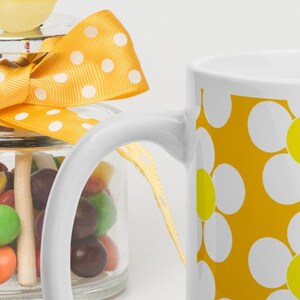 May include: A white ceramic mug with a yellow background and white daisy pattern. The mug is next to a glass jar filled with colorful candies and a yellow ribbon.