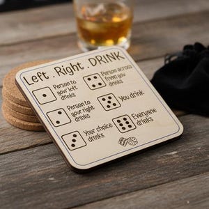 Könnte beinhalten: Ein Getränkeuntersetzer aus Holz mit dem Text "Left. Right. DRINK" und Würfelanweisungen. Der Untersetzer ist hellbraun mit einem dunkelbraunen Rand. Ein Stapel Untersetzer aus Kork und ein Glas bernsteinfarbener Flüssigkeit befinden sich im Hintergrund.