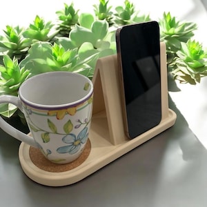 May include: A beige phone stand with a smartphone, a floral-decorated mug, and a cork coaster. The stand is on a tray with a curved edge. Green succulents are in the background.