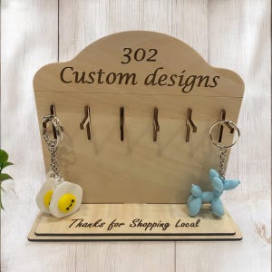 May include: Wooden key rack with five hooks and the text "302 Custom designs" and "Thanks for Shopping Local". Two keychains are hanging from the hooks, one with a blue balloon dog and the other with a white egg with a yellow yolk.