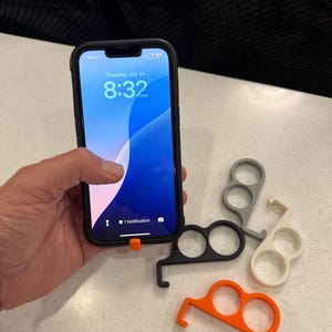 May include: A black smartphone in a case displays the time and date. Several touch-free door opener tools in black, gray, white, and orange are arranged on a white surface. The tools have finger holes and a hook.