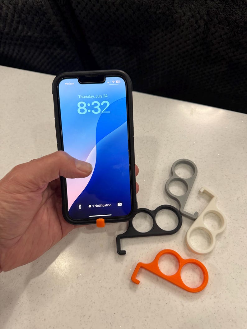 May include: A black smartphone in a case displays the time and date. Several touch-free door opener tools in black, gray, white, and orange are arranged on a white surface. The tools have finger holes and a hook.