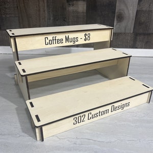 K&ouml;nnte beinhalten: Ein dreistufiger Holzst&auml;nder mit schwarzem Besatz. Die oberste Stufe tr&auml;gt den Text "Coffee Mugs - $8". Die unterste Stufe tr&auml;gt den Text "302 Custom Designs".