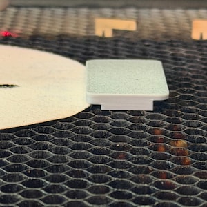 May include: A small, light green, square-shaped object with a textured surface and a white base. The object is likely a component or part of a larger item, possibly for crafting or assembly. The background features a dark, honeycomb-patterned surface.