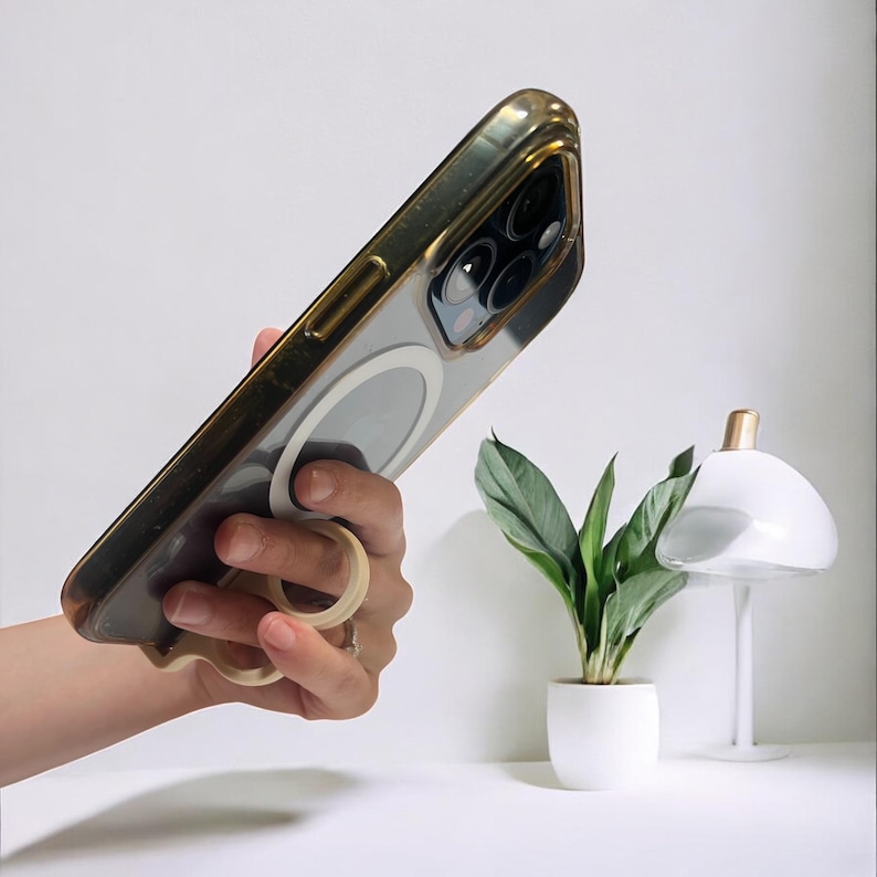 May include: Clear phone case with a gold-colored frame and a ring holder. The phone case is held up, showing the camera lenses. A small white lamp with a gold top and a potted plant are in the background.