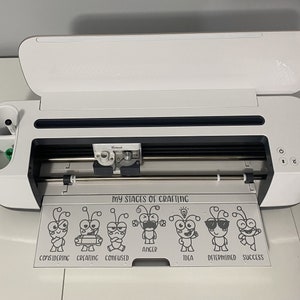 Pu&ograve; includere: Una macchina da taglio Cricut Explore Air 2 bianca con il coperchio aperto. La macchina &egrave; su una superficie bianca e ha un tappetino da taglio nero con un design che dice "Le mie fasi del crafting" con una serie di insetti dei cartoni animati che rappresentano diverse fasi del crafting. Gli insetti sono etichettati come "Considerando", "Creando", "Confuso", "Rabbia", "Idea", "Determinato" e "Successo".
