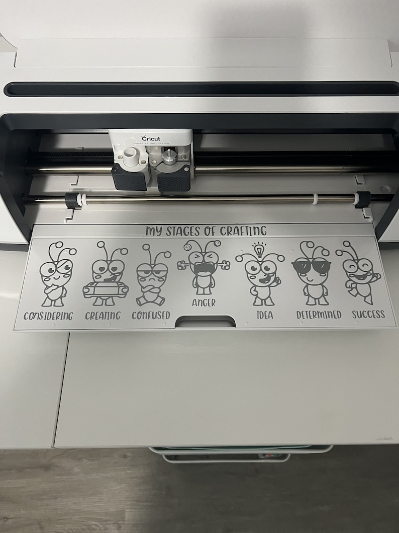 Pu&ograve; includere: Una macchina da taglio Cricut bianca con un design in bianco e nero sul tappetino che dice "Le mie fasi dell'artigianato" con una serie di formiche dei cartoni animati che rappresentano diverse fasi dell'artigianato: considerando, creando, confuso, rabbia, idea, determinato e successo.