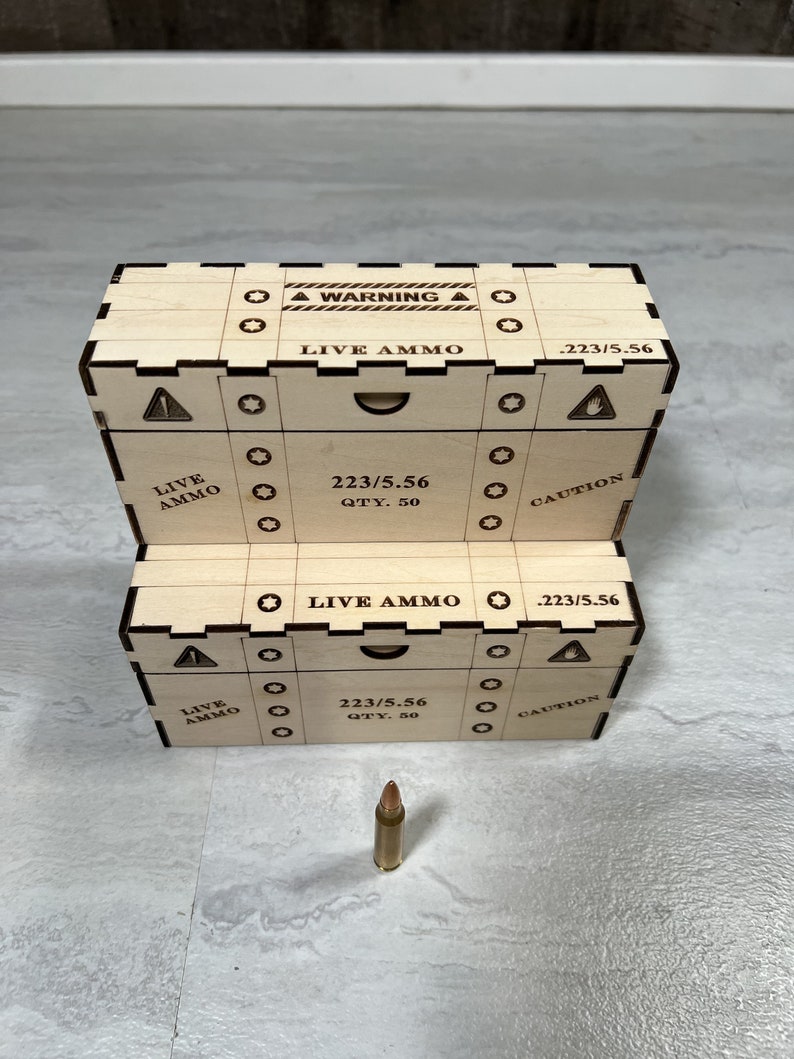 K&ouml;nnte beinhalten: Holzkiste mit einem Warnhinweis mit der Aufschrift "WARNING LIVE AMMO" und einem Etikett mit der Aufschrift "223/5.56". Die Kiste hat eine Schublade mit der Aufschrift "LIVE AMMO" und einem Etikett mit der Aufschrift "223/5.56 QTY. 50 CAUTION". Eine einzelne Kugel befindet sich vor der Kiste.