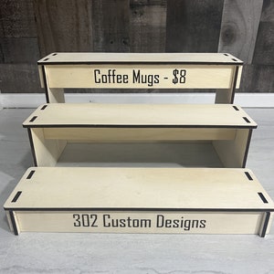 K&ouml;nnte beinhalten: Ein dreistufiger Holzst&auml;nder mit heller Holzoptik. Die oberste Stufe tr&auml;gt den Text "Coffee Mugs - $8" und die unterste Stufe den Text "302 Custom Designs".