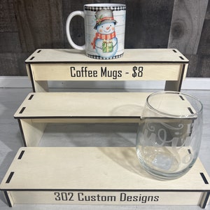 K&ouml;nnte beinhalten: Eine wei&szlig;e Kaffeetasse mit einem Schneemann-Design und einem schwarz-wei&szlig; karierten Rand. Die Tasse steht auf einem Holzregal mit dem Text "Coffee Mugs - 8 &euro;".