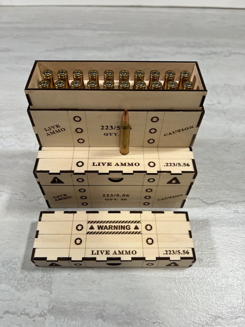 K&ouml;nnte beinhalten: Eine Holzkiste mit einem Warnhinweis, auf dem "WARNING" und "LIVE AMMO" steht. Die Kiste ist ge&ouml;ffnet und enth&auml;lt eine Reihe goldfarbener Kugeln. Die Kiste tr&auml;gt auch ein Etikett mit der Aufschrift "223/5.56".