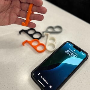 May include: An orange, plastic, no-touch door opener and stylus held in a hand, with several others in black, white, and gray. A smartphone with a black case is also in the image, displaying the time 8:36.