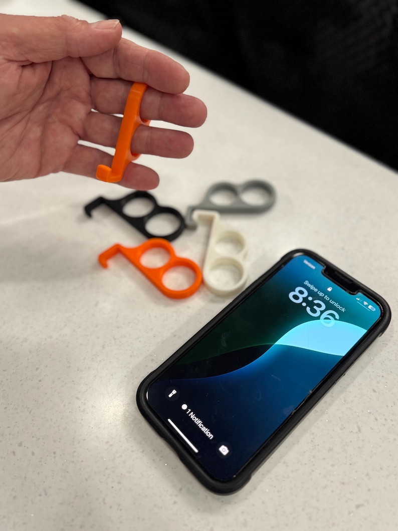 May include: An orange, plastic, no-touch door opener and stylus held in a hand, with several others in black, white, and gray. A smartphone with a black case is also in the image, displaying the time 8:36.