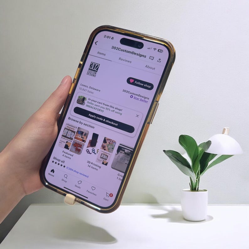 May include: A smartphone with a gold-colored case displays an Etsy shop page. The screen shows the shop name "302CustomDesigns" and various product images. A small potted plant with green leaves and a white pot is next to the phone.
