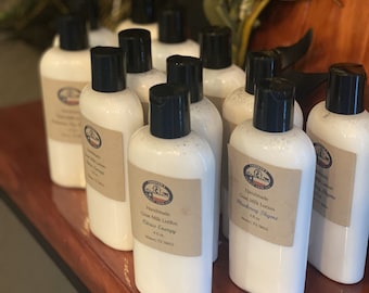 May include: A collection of white bottles with black lids and labels. The labels have a rustic design and list the product as "Handmade Goat Milk Lotion" with different scents, including "Citrus Energy" and "Blueberry Thyme".