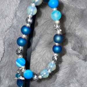 May include: A beaded bracelet featuring a mix of blue and clear beads. The bracelet includes various shades of blue beads, including solid and patterned designs, interspersed with silver-colored spacers and a small charm.