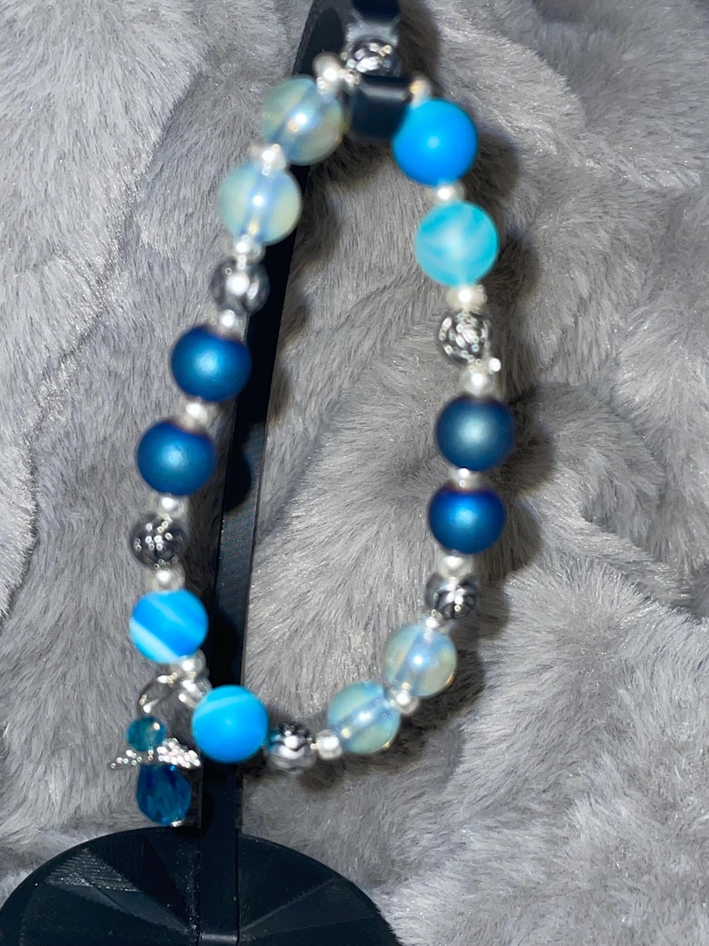 May include: A beaded bracelet featuring a mix of blue and clear beads. The bracelet includes various shades of blue beads, including solid and patterned designs, interspersed with silver-colored spacers and a small charm.