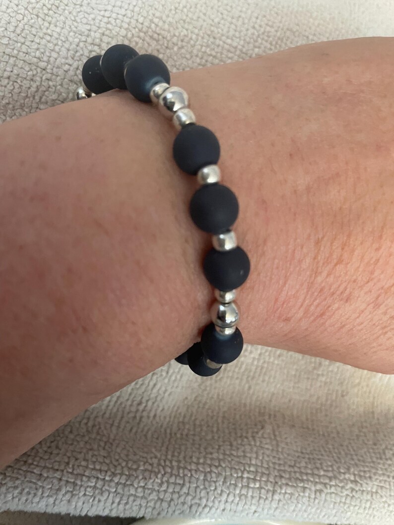 May include: A dark blue beaded bracelet with silver-colored metal accents. The bracelet features matte, round beads interspersed with smaller, shiny metal beads. The bracelet is worn on a wrist.