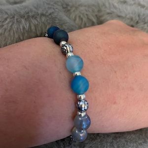 May include: A beaded bracelet featuring a gradient of blue and gray beads. The bracelet has a mix of matte and shiny beads, with silver-colored decorative spacers. The bracelet is on a person's wrist.