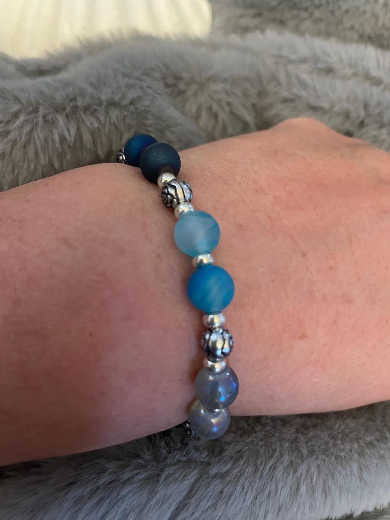 May include: A beaded bracelet featuring a gradient of blue and gray beads. The bracelet has a mix of matte and shiny beads, with silver-colored decorative spacers. The bracelet is on a person's wrist.