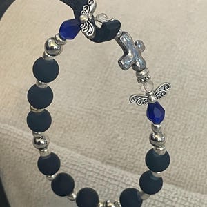 May include: A beaded bracelet featuring matte black beads, silver-colored accents, and decorative charms. The bracelet includes a cross charm, blue crystal accents, and angel-shaped embellishments. The bracelet is displayed on a black leather strap.