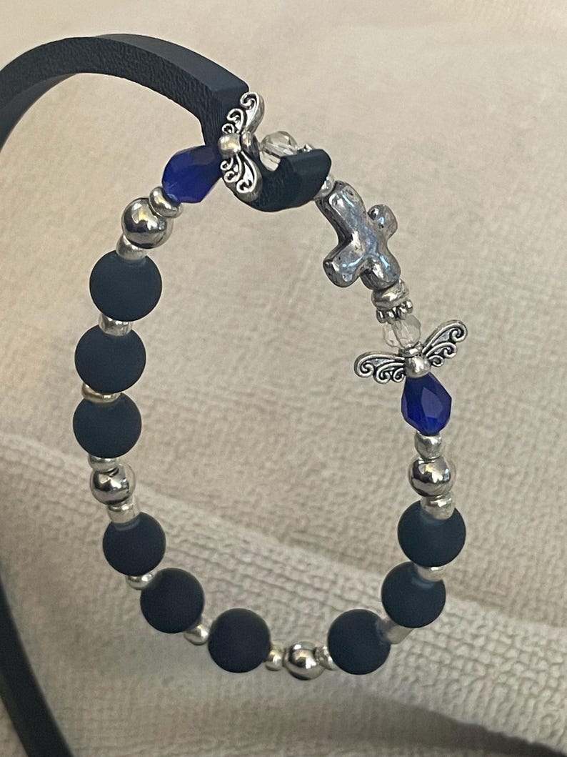 May include: A beaded bracelet featuring matte black beads, silver-colored accents, and decorative charms. The bracelet includes a cross charm, blue crystal accents, and angel-shaped embellishments. The bracelet is displayed on a black leather strap.