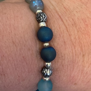 May include: A beaded bracelet featuring a mix of blue and silver beads. The bracelet includes matte blue and iridescent beads, silver rose-shaped accents, and small silver spacer beads. The bracelet is strung on an elastic cord.