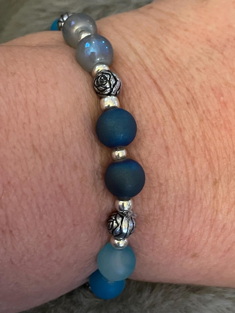 May include: A beaded bracelet featuring a mix of blue and silver beads. The bracelet includes matte blue and iridescent beads, silver rose-shaped accents, and small silver spacer beads. The bracelet is strung on an elastic cord.